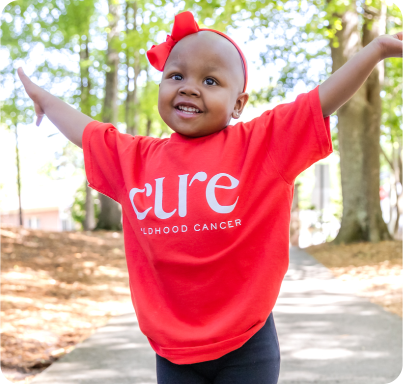 Young childhood cancer patient wearing red CURE t-shirt with arms raised smiling representing hope and success in pediatric cancer research and treatment