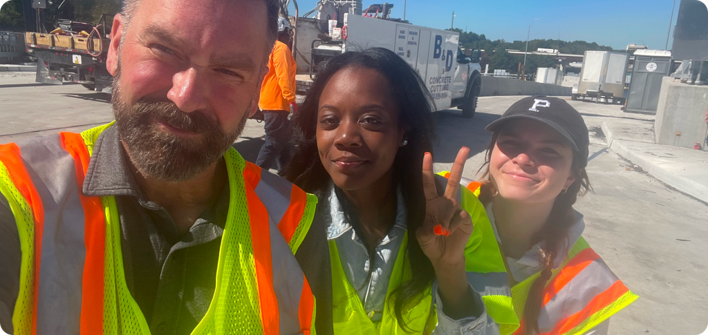 Three individuals wearing safety vests posing for a selfie at a worksite.