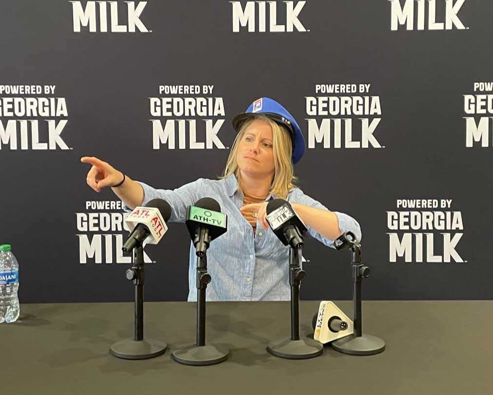 Woman with a hat on in front of four microphones pretending to take questions
